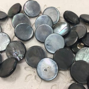 Peut inclure: Un ensemble de boutons ronds gris foncé avec un éclat nacré. Les boutons sont de tailles variables et ont une forme plate en forme de disque. Certains boutons présentent une qualité irisée subtile, reflétant la lumière. Un dé à coudre est visible en arrière-plan.