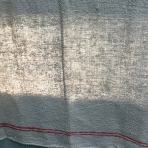 May include: A close-up of a light gray linen fabric with two red stripes along the bottom edge. The fabric has a textured, woven appearance and is slightly wrinkled.