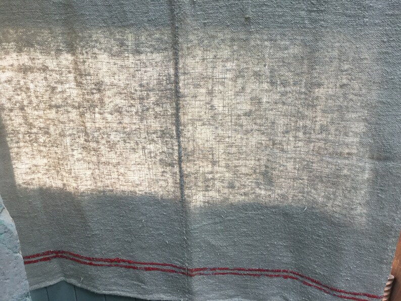 May include: A close-up of a light gray linen fabric with two red stripes along the bottom edge. The fabric has a textured, woven appearance and is slightly wrinkled.