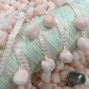 Peut inclure: Une bobine de fil blanc avec une bordure de pompons roses. Le fil est enroulé autour d'un tube en carton. Les pompons sont doux et moelleux.
