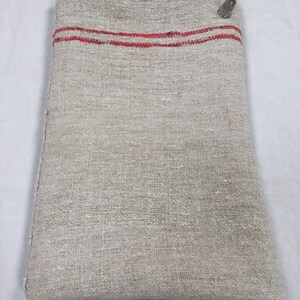 May include: A folded linen fabric with two red stripes along the top edge. The fabric is a natural off-white color and has a rough texture.