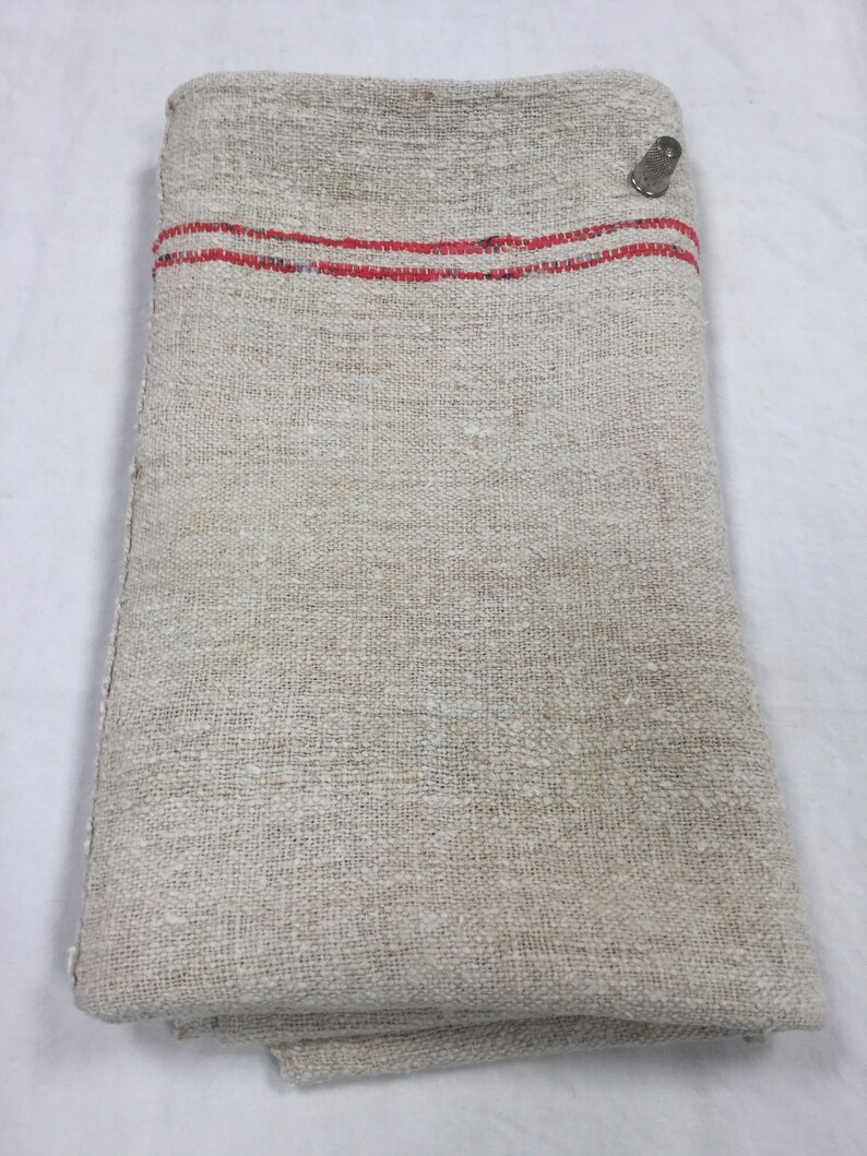 May include: A folded linen fabric with two red stripes along the top edge. The fabric is a natural off-white color and has a rough texture.