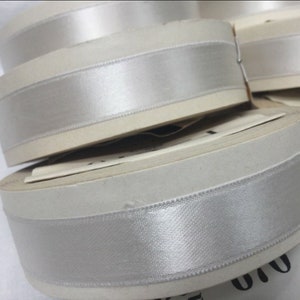 Puede incluir: Rollos de cinta de satén blanco con una raya plateada. La cinta está enrollada alrededor de carretes de cartón.