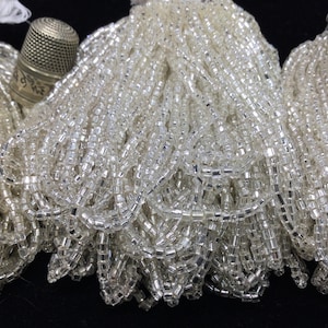 Peut inclure: Gros plan de plusieurs brins de perles rectangulaires transparentes. Un dé à coudre argenté repose parmi les perles. Les perles sont enfilées ensemble, créant un effet chatoyant. Les perles sont probablement utilisées pour la fabrication de bijoux ou d'autres travaux manuels.