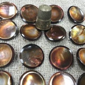 Puede incluir: Una colección de botones redondos iridiscentes con bordes marrón oscuro. Un dedal plateado está centrado entre los botones. Los botones tienen una calidad brillante y nacarada, con colores que van del dorado al marrón. Los botones están dispuestos sobre una superficie clara y texturizada.