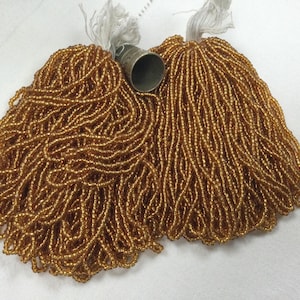 Puede incluir: Dos paquetes de cuentas de semillas doradas con borlas de hilo blanco. Una dedal plateado se coloca entre los paquetes. Las cuentas están fuertemente ensartadas, creando una apariencia texturizada y fluida. Ideal para la fabricación de joyas.