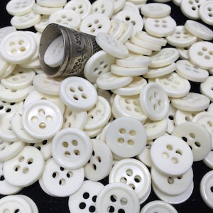 May include: A large pile of white plastic buttons with four holes each. The buttons are arranged in a random pattern and are all the same size. There is a small silver thimble in the pile of buttons.