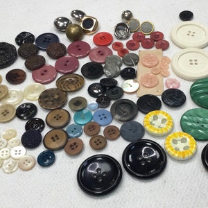 Op de afbeelding: Een verzameling diverse knopen in verschillende maten, kleuren en materialen. De collectie bevat knopen in bruin-, zwart-, wit-, rood- en blauwtinten. Sommige knopen hebben vier gaten, andere twee of zijn schachtknopen. Er is ook een vingerhoed.