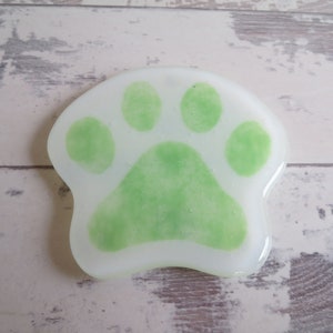 May include: A white glass coaster with a green paw print design. The paw print has four toes and a pad.