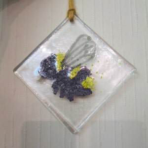 Può includere: Un ornamento di vetro trasparente a forma di diamante con un'immagine dipinta di un'ape con accenti gialli e viola. L'ornamento è appeso a una corda dorata.