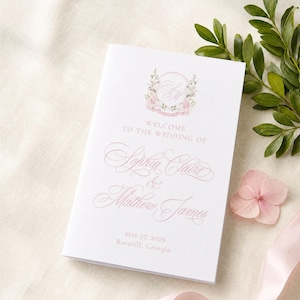 Könnte beinhalten: Ein weißes Hochzeitsheft mit einem rosa Blumenkranz-Monogramm oben. Der Text lautet: Welcome to the wedding of Sophia Claire & Mathew James, May 27, 2029, Roswell, Georgia, gedruckt in eleganten rosa Schreib- und Serifenschriften.