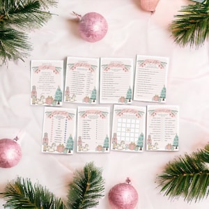 May include: A collection of baby shower game cards with a Christmas theme. The cards feature illustrations of nutcrackers, Christmas trees, and gift boxes in pastel colors. Pink ornaments and evergreen branches surround the cards.