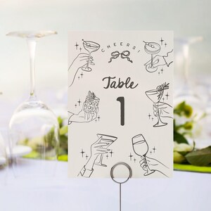 May include: A white table number card with black ink. The card has a black and white illustration of a cocktail glass and the text "Cheers!" and "Table 1".