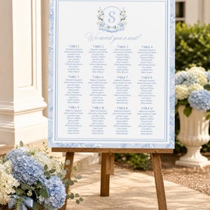 May include: A rectangular wedding seating chart sign on a wooden easel. The sign features a light blue floral border, the text "We saved you a seat!", a central monogram "S" inside a wreath, and a list of guests organized by twelve tables.