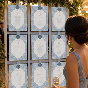 May include: A wedding seating chart display featuring nine rectangular cards arranged in a grid on a wooden frame. Each card has a light blue floral pattern, a central white panel with table numbers one through nine, and a blue wax seal at the top.