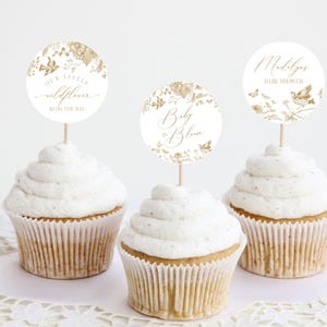 Puede incluir: Tres cupcakes con glaseado blanco y adornos decorativos. Los adornos son círculos blancos con diseños florales dorados y texto que dice "Our Little Wildflower is on the Way", "Baby is Bloom" y "Madalyn's Baby Shower".