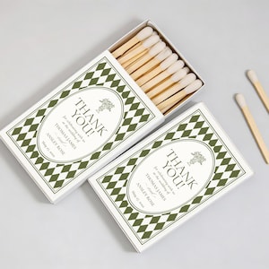 May include: Two white matchboxes with a green diamond pattern and the words "THANK YOU!" printed on them. The boxes contain wooden matches with white tips. The text also includes names and a date.