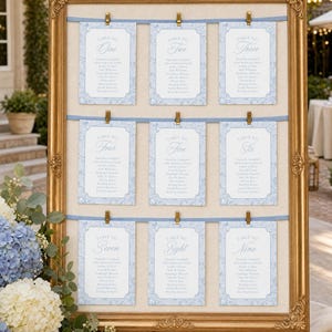 May include: A gold ornate frame holds a seating chart with nine light blue cards clipped to horizontal ribbons. Each card features a table number from one to nine in script text, followed by a list of names printed in a dark blue font.