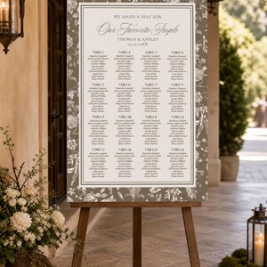 Puede incluir: Un cartel rectangular de distribución de asientos para bodas con un borde color topo y un patrón floral blanco. El centro es blanco con el texto WE SAVED A SEAT FOR Our Favorite People, THOMAS & ANSLEY, 03.23.20XX, y dieciséis mesas con nombres.