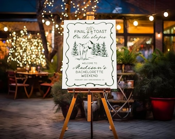 May include: A white sign on a wooden easel with the text "FINAL TOAST On the slopes" and "WELCOME TO Madison's BACHELORETTE WEEKEND". The sign has a green border and a mountain and tree illustration. The background is an outdoor setting with string lights.