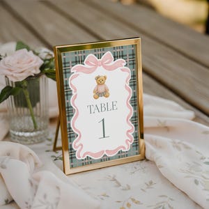 May include: A gold-framed table number card with a plaid background and a pink bow design. The card features a teddy bear illustration and the number 1. The card is displayed on a wooden table with floral accents.