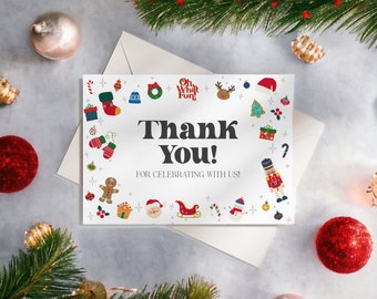 May include: A white greeting card with the words "Thank You!" and "For Celebrating With Us!" surrounded by Christmas-themed illustrations. The card is on a white envelope, with Christmas ornaments and greenery in the background.