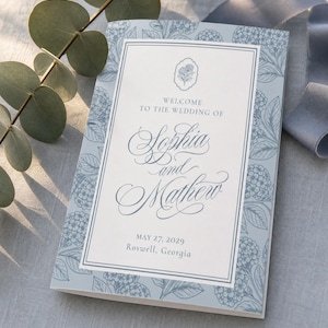 Pode incluir: Um programa de casamento retangular com uma borda azul-clara e uma ilustração botânica de hortênsias em azul-escuro. O painel central branco exibe o texto: Welcome to the wedding of Sophia and Matthew, May 27, 2029, Roswell, Georgia.