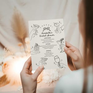 May include: A black and white printable bridal shower menu with the title "Annabelle's Bridal Shower" and a list of appetisers, entrees, and desserts. The menu is decorated with hand-drawn illustrations of cocktails.