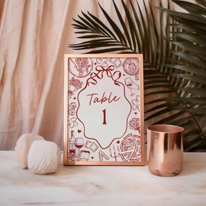 Puede incluir: Un marco de metal color cobre contiene un letrero blanco con las palabras "Table 1" en una fuente decorativa. El letrero está rodeado de ilustraciones rojas de comida y bebida. También hay una taza de metal color cobre y dos esferas decorativas blancas.