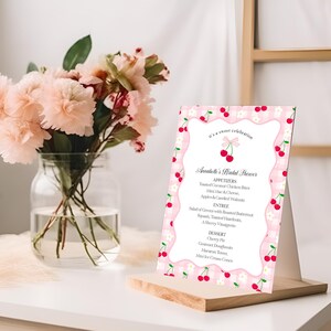 May include: A pink and white checkered menu card with a cherry design, displayed on a wooden stand. The menu lists appetizers, entrees, and desserts for Annabelle's Bridal Shower. A vase of pink flowers sits to the left.