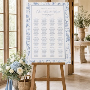May include: A rectangular wedding seating chart sign on a wooden easel, featuring a blue and white floral border. The text reads: WE SAVED A SEAT FOR Our Favorite People, THOMAS & ANSLEY, 03.23.20XX, followed by sixteen tables of guest names.