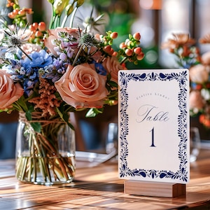 May include: A table number card with a white background and a navy blue floral border. The card reads "Cielito Lindo" and "Table 1" in elegant script. The card is displayed on a wooden block, with a bouquet of flowers in the background.