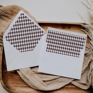 May include: Two white envelopes with a brown diamond pattern on the inside. The envelopes are open, revealing the patterned interior. They are arranged on a wooden tray with a tan fabric.