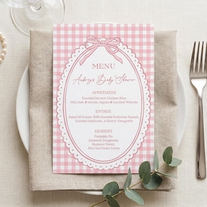 May include: A pink and white gingham baby shower menu with the text "Aubrey's Baby Shower" in elegant script. The menu includes appetizers, entree, and dessert options. A pearl necklace, silverware, and greenery complete the table setting.