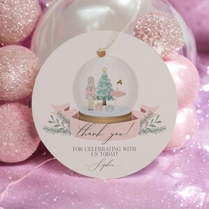 May include: A round, light pink gift tag with a snow globe illustration featuring a nutcracker, ballerina, and Christmas tree. The tag includes the text "thank you!" and "For celebrating with us today!" with a name at the bottom.