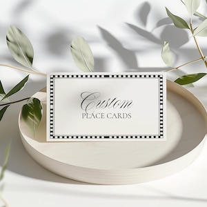 May include: A rectangular place card with the words "Custom PLACE CARDS" in elegant script. The card has a black and white border and sits on a light-colored, round wooden tray. Green leaves and branches are in the background.