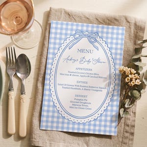 May include: A baby shower menu with a blue and white gingham pattern. The menu features an oval frame with a bow at the top and the text "Aubrey's Baby Shower." Silverware and a glass of wine are also visible.