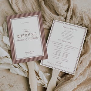 May include: Two wedding fan programs with wooden handles. The fans have a white background with text in a burgundy frame. One fan says "Welcome to The Wedding Thomas & Ansley" and the other lists the order of events and wedding party.