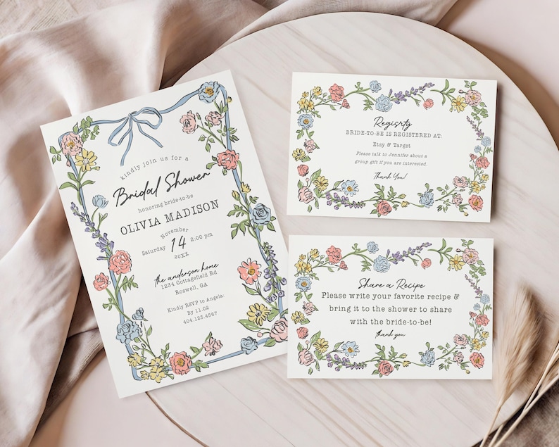 Może przedstawiać: Zaproszenia i karty na baby shower z kwiatowymi obramowaniami. Gł&oacute;wne zaproszenie zawiera niebieską wstążkę i tekst, w tym imię panny młodej i szczeg&oacute;ły wydarzenia. Dodatkowe karty zawierają informacje.