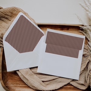 Peut inclure: Deux enveloppes blanches avec un intérieur à rayures marron, dont une ouverte et une avec une carte à l'intérieur, reposent sur un plateau en bois. Les enveloppes et la carte sont posées sur un fond neutre avec un tissu drapé.