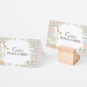 May include: Two place cards with a light brown gingham background and a floral wreath. The wreath has a white goose with a pink and white hat inside. The text on the cards reads "Custom Place Cards".
