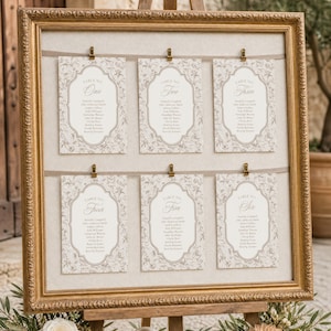 Puede incluir: Un marco dorado ornamentado que sostiene un plano de asientos con seis tarjetas rectangulares sujetas a dos cintas horizontales. Cada tarjeta presenta un borde floral, el texto Table No, un número del One al Six y una lista de nombres.