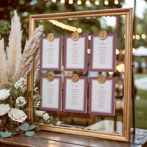 Peut inclure: Un miroir doré encadre six cartons de table pour un mariage. Chaque carton a un fond blanc avec une bordure rayée bordeaux et un sceau de cire doré. Les cartons sont numérotés de un à six.