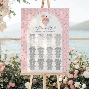 May include: A pink and white bridal shower seating chart on an easel. The chart features a colorful ice cream sundae graphic and the text "Take a Seat" and "Sophia's Bridal Shower." The chart lists guest names under table numbers.