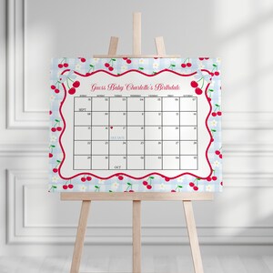 May include: A baby shower calendar on an easel. The calendar has a light blue gingham background with a red cherry border. The text "Guess Baby Charlotte's Birthdate" is at the top. The calendar grid is for September and October.