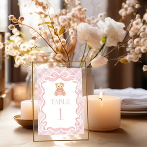 May include: A table number card in a gold frame, featuring a pink plaid design and a teddy bear illustration. The card displays "TABLE 1". The scene includes candles and floral arrangements. The card is likely for a wedding or event.