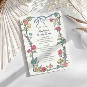 Puede incluir: Un menú floral para una despedida de soltera sobre papel blanco. El menú presenta una cinta azul y un borde de flores rosas, amarillas y azules. El texto del menú dice "LET'S EAT" y "Madison's Bridal Shower".