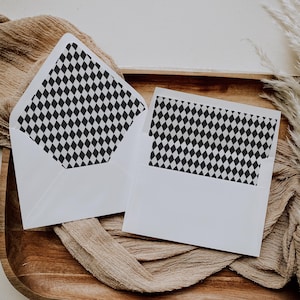 May include: Two white envelopes with a black and white diamond pattern lining. The envelopes are open, revealing the patterned interior. They rest on a wooden tray with a tan fabric and dried plants.