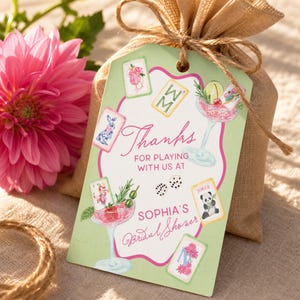 May include: A gift tag with a light green background and pink accents, featuring the text "Thanks for playing with us at Sophia's Bridal Shower." The tag is adorned with playing card illustrations and a cocktail glass design. The tag is attached to a burlap bag.