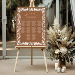 May include: A brown wedding seating chart with a white floral border. The chart is on an easel and says "Take a Seat" with the names of the bride and groom.